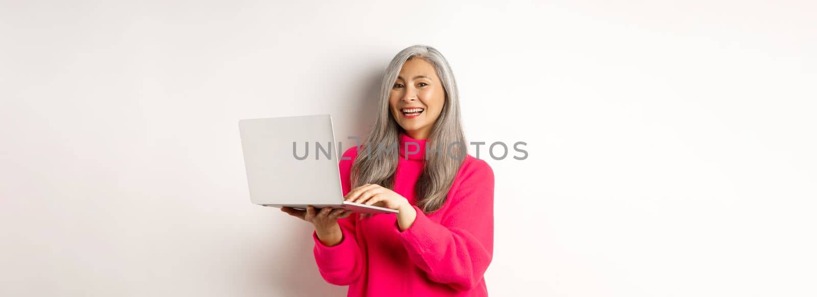 Beautiful asian senior woman entrepreneur working with laptop, laughing and smiling at camera, standing over white background.