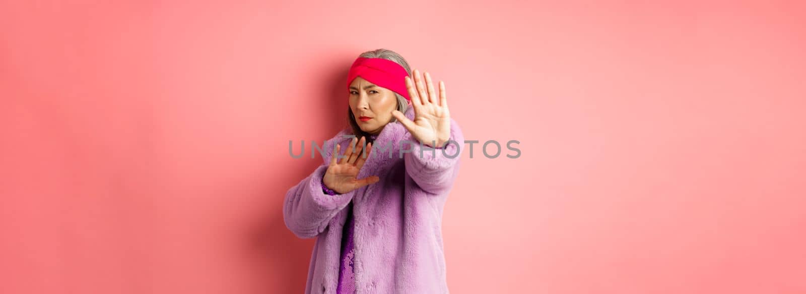 Asian senior female model in stylish purple winter coat standing in victim pose, extending hand in stop gesture and pleading for mercy, standing against pink background.
