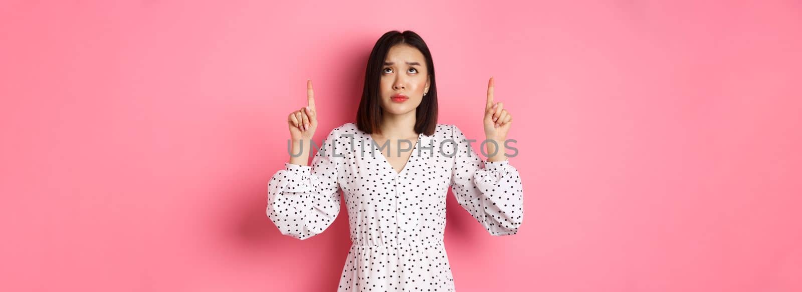 Worried and concerned asian girl in cute dress frowning, pointing and looking up with sad face, standing over pink background. Copy space