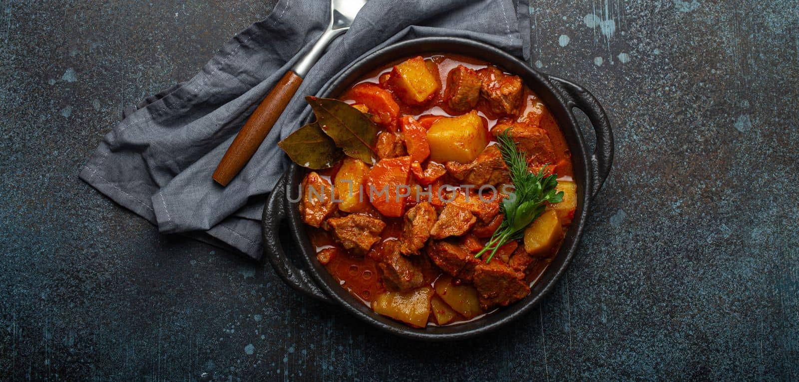Beef meat stew with potatoes, carrot and delicious gravy in black casserole pot with bay leaves and fresh green herbs with spoon on black dark rustic concrete background from above . by its_al_dente