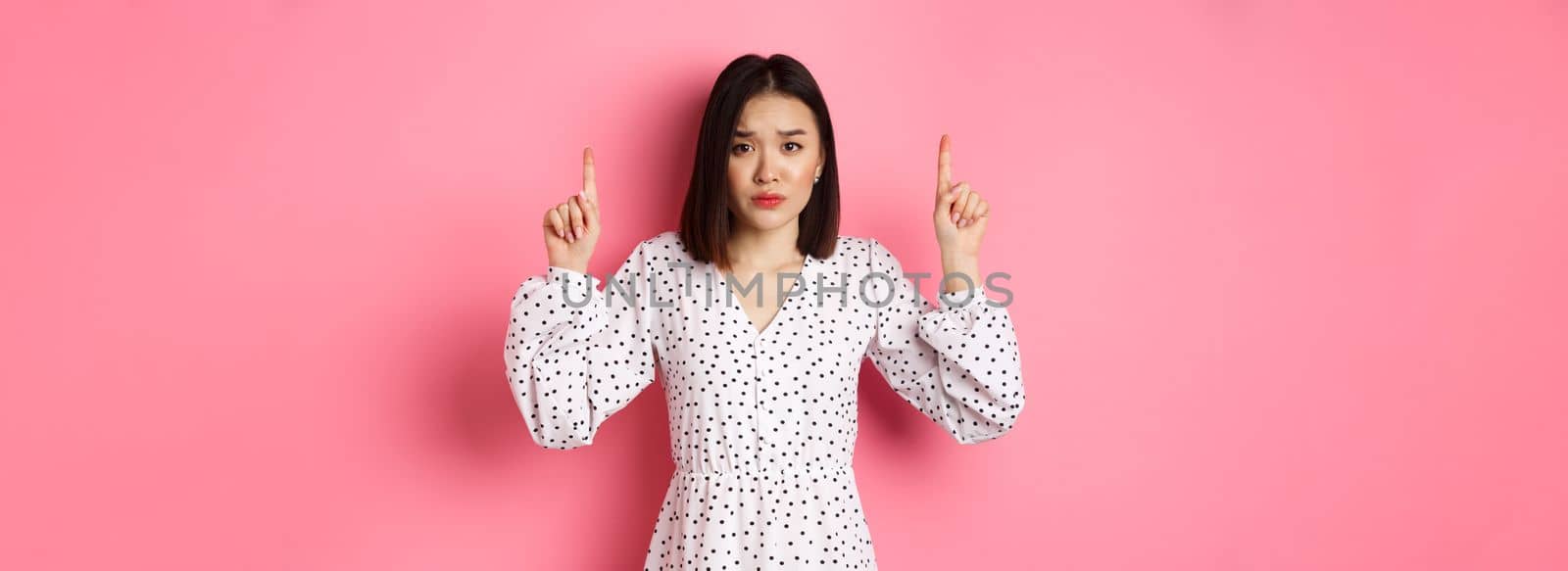 Upset asian girl pointing fingers up, showing promo banner, staring at camera with disappointed gloomy look, standing in dress over pink background by Benzoix