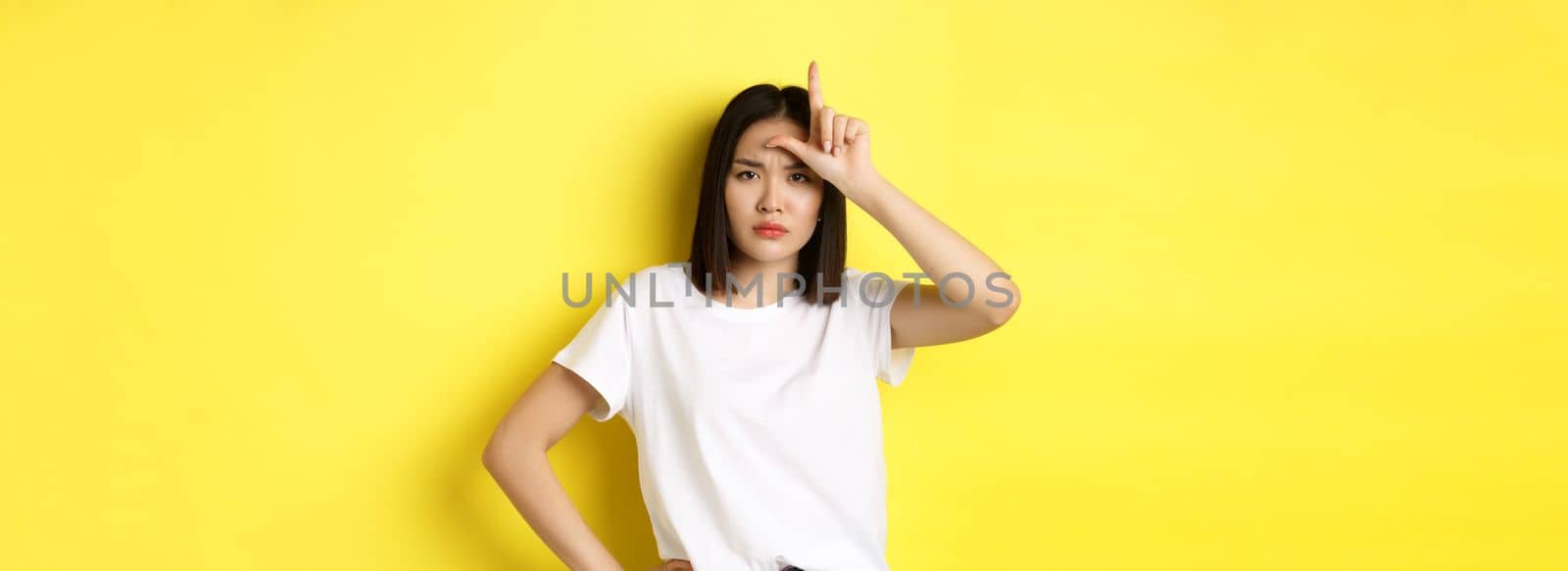 Serious and arrogant asian woman mocking lost team, showing loser sign on forehead and stare at camera confident, standing over yellow background.