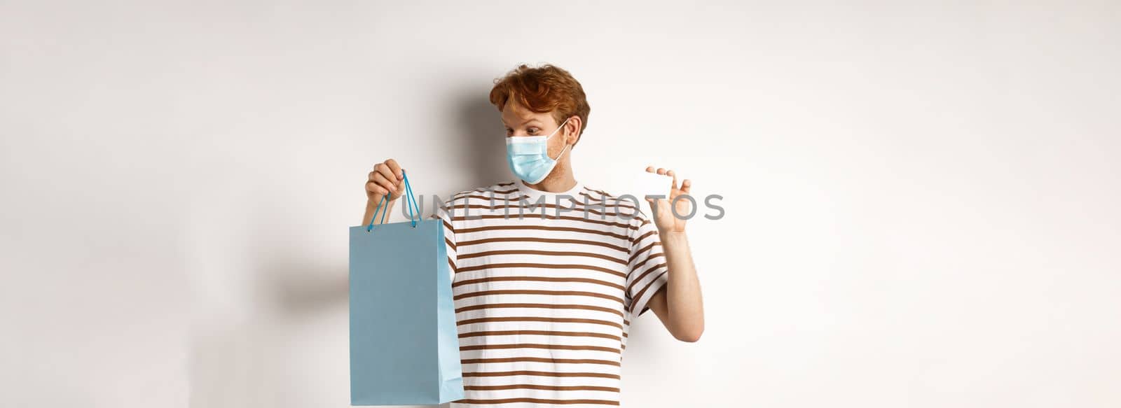 Concept of covid-19 and lifestyle. Cheerful young man with red hair, wear medical mask, showing shopping bag from store and plastic credit card.