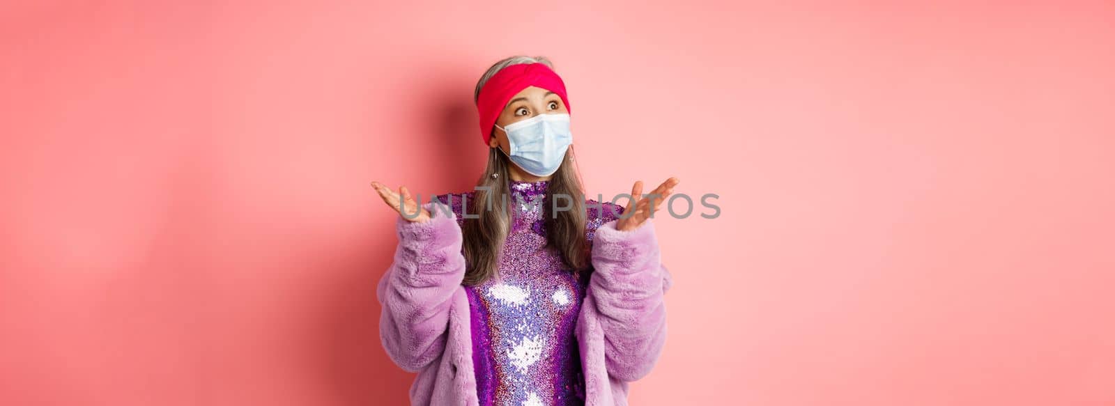 Covid-19, social distancing and fashion concept. Surprised and shocked asian stylish granny looking left, spread hands sideways confused, standing over pink background.