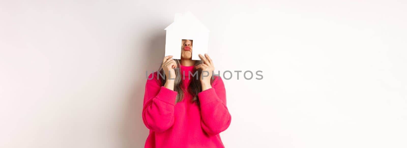 Real estate. Funny asian middle-aged woman hiding face behind paper house model and showing puckered lips, having fun, standing over white background.