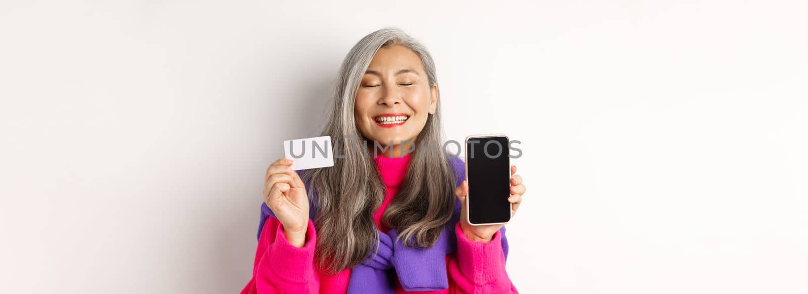 Online shopping. Happy and delighted asian senior woman smiling with joy, showing blank smartphone screen and plastic card, buying in internet, white background.