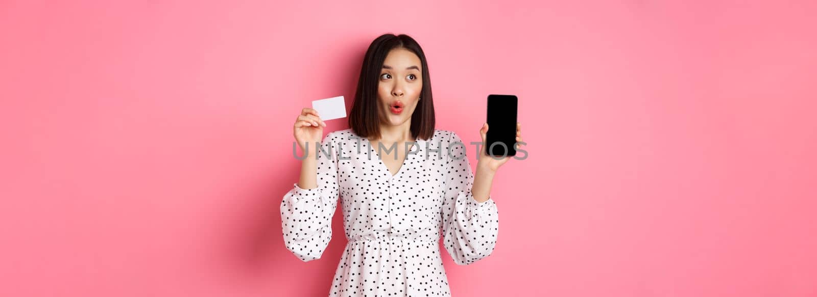 Cute asian woman shopping online, showing bank credit card and mobile screen, smiling and looking left at copy space, standing over pink background by Benzoix