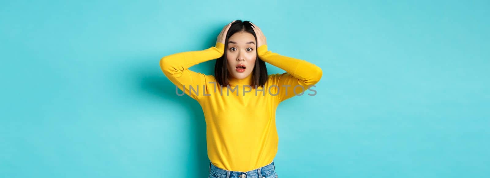 Shocked asian woman looking speechless, gasping amazed and holding hands on head, hear big news, standing over blue background.