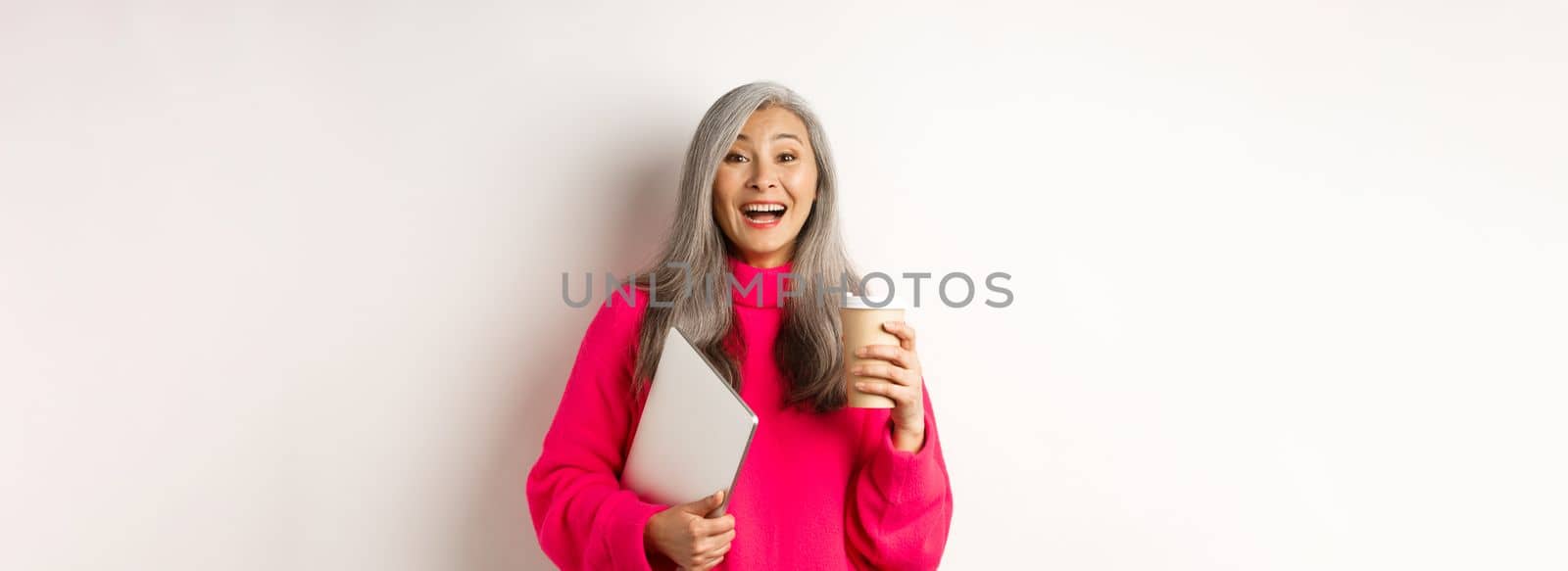 Happy asian senior female entrepreneur laughing, drinking coffee and holding laptop, standing in pink sweater over white background.