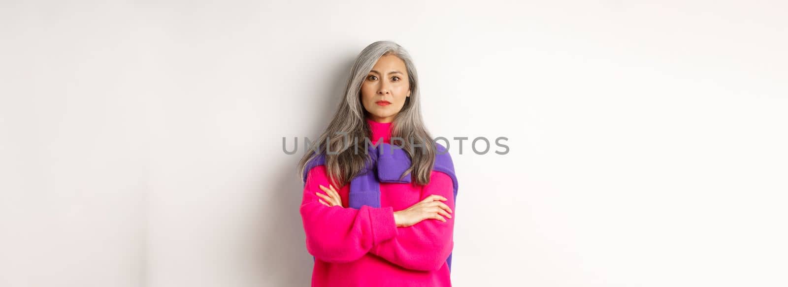 Serious and strict asian grandmother looking angry at camera, cross arms on chest, standing over white background disappointed.