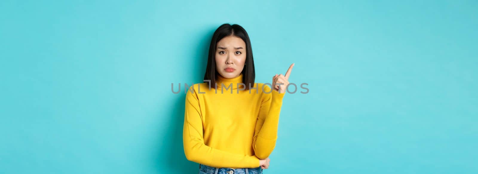 Shopping concept. Sad and gloomy asian girl in yellow sweater pointing finger left, frowning and showing bad news on copy space, standing over blue background by Benzoix