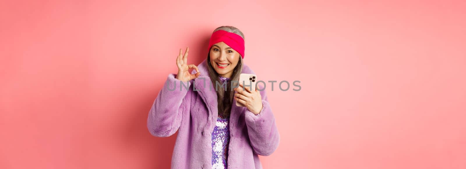 Online shopping and fashion concept. Stylish asian senior woman showing okay sign and holding mobile phone, recommending internet store, pink background.
