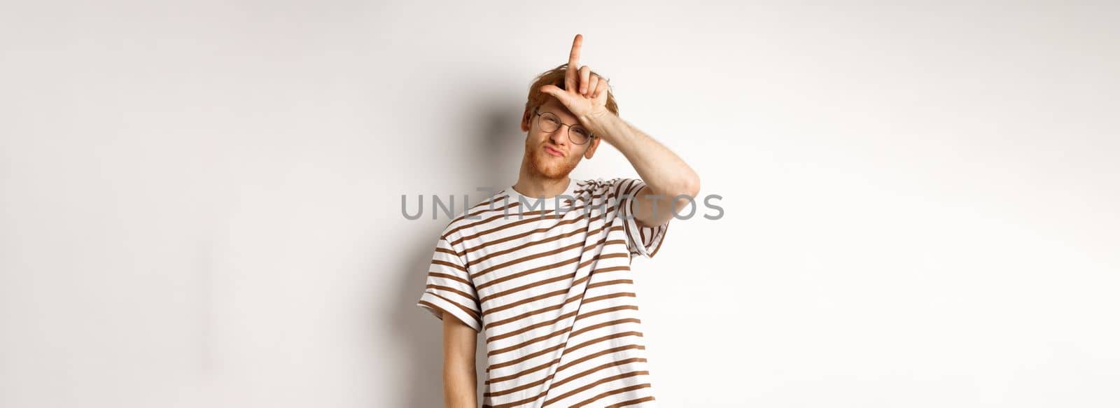 Sassy and arrogant redhead man scolding lost team, showing loser gesture on forehead and making smug face, standing over white background.