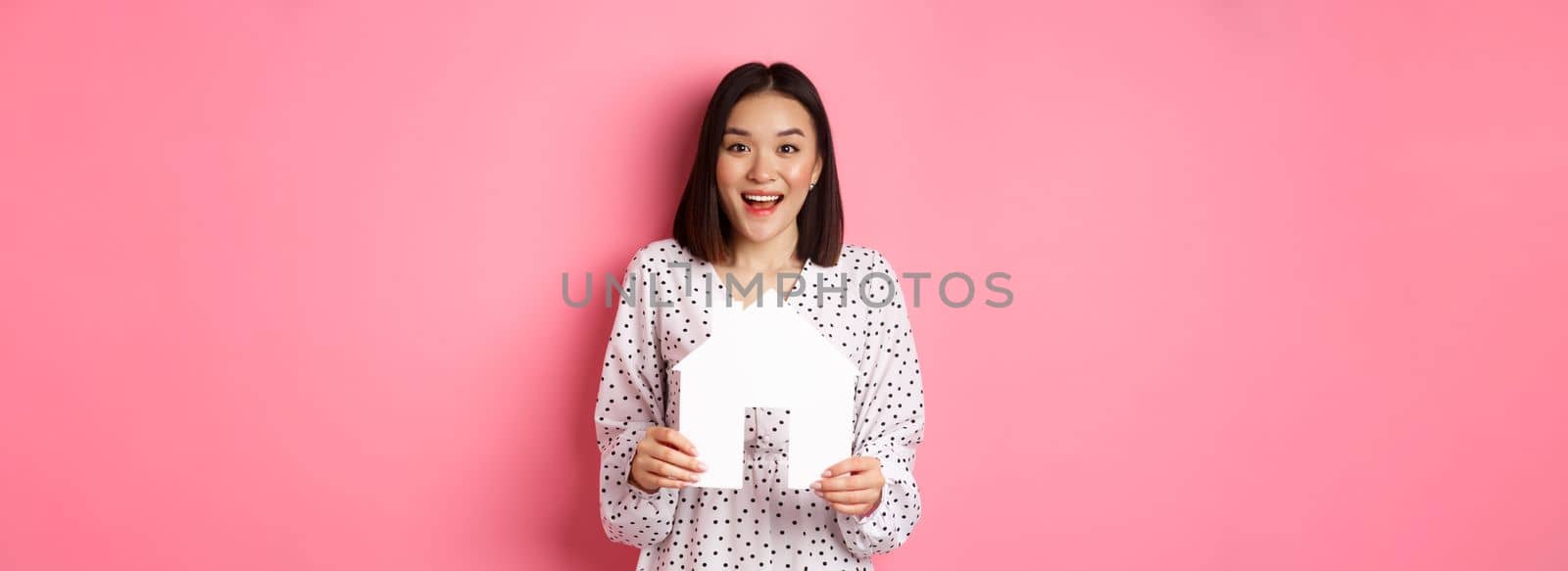 Real estate. Amazed asian woman searching for flat, holding house model and looking at camera happy, standing over pink background.