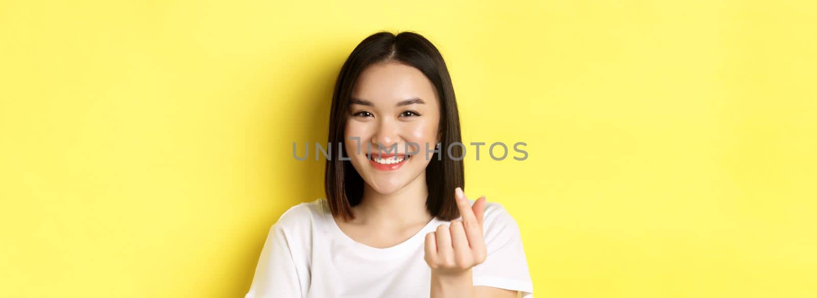 Valentines day and women concept. Close up of pretty asian girl in white t-shirt, smiling and showing finger heart, standing over yellow background.