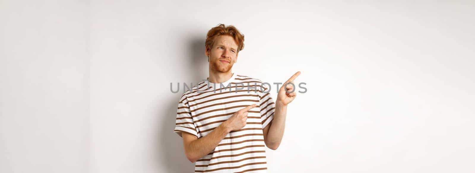 Doubtful young man with bristle and red hair looking, pointing fingers left at something average, staring skeptical at promo, white background.