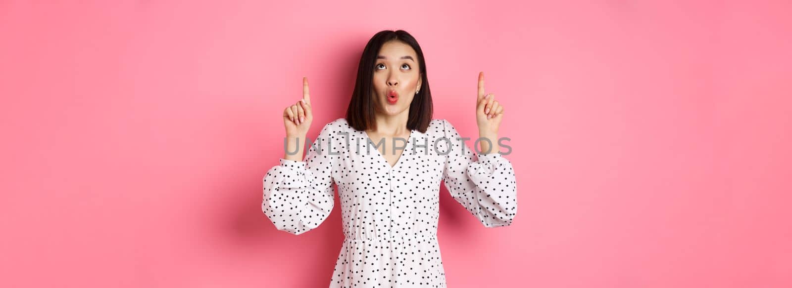 Cute korean girl in beautiful dress saying wow, looking and pointing fingers up, intrigued in promo offer, standing over pink background by Benzoix