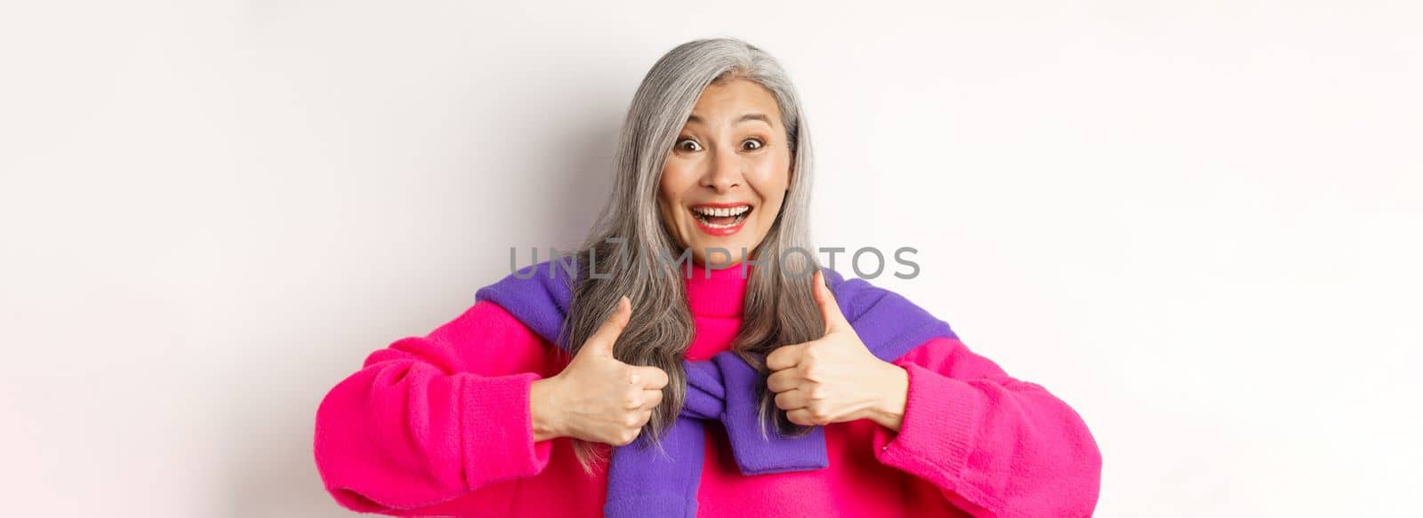 Excited and amazed asian elderly woman showing thumbs-up in approval, smiling happy at camera, praising something awesome, white background.