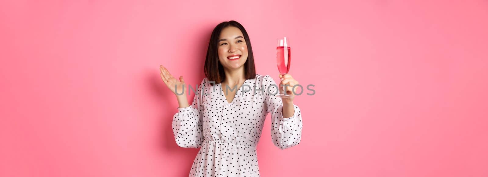 Beautiful asian woman, raising glass of champagne and smiling happy, drinking wine and celebrating, standing over pink background by Benzoix