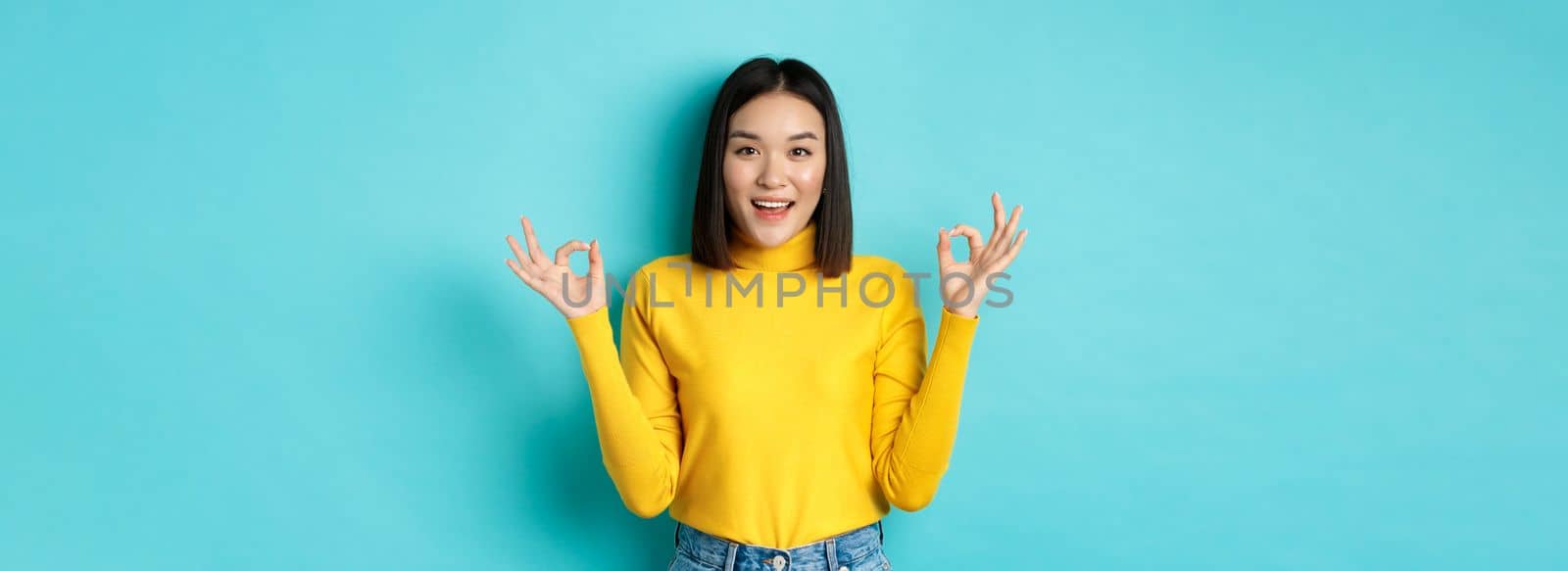 Cheerful asian female model showing okay gestures, smiling and looking impressed, praise product, standing over blue background.