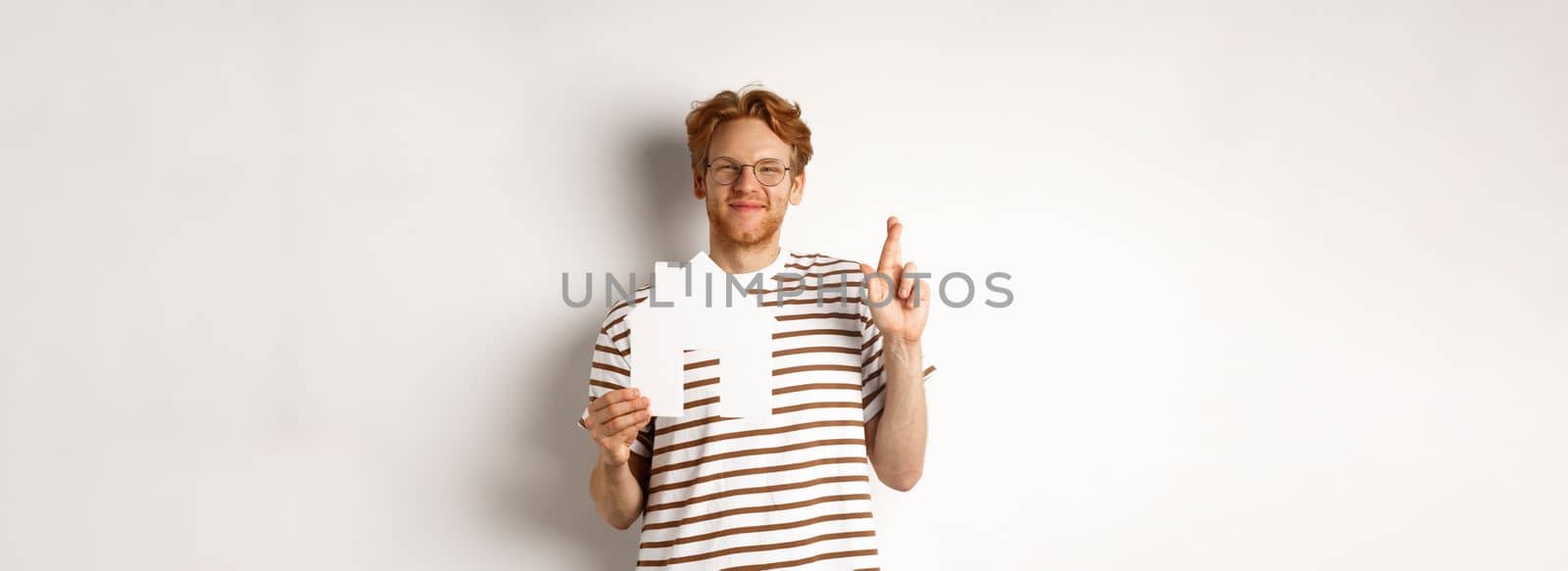 Hopeful redhead man dreaming of buying house, holding paper home cutout and cross fingers for good luck, making a wish, standing over white background.