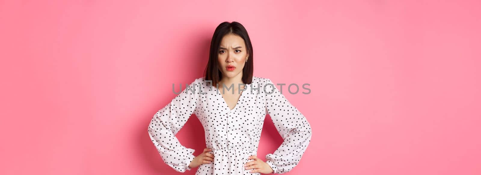 Angry asian woman staring at camera frustrated, holding hands on waist and waiting for explanations, standing in dress against pink background.