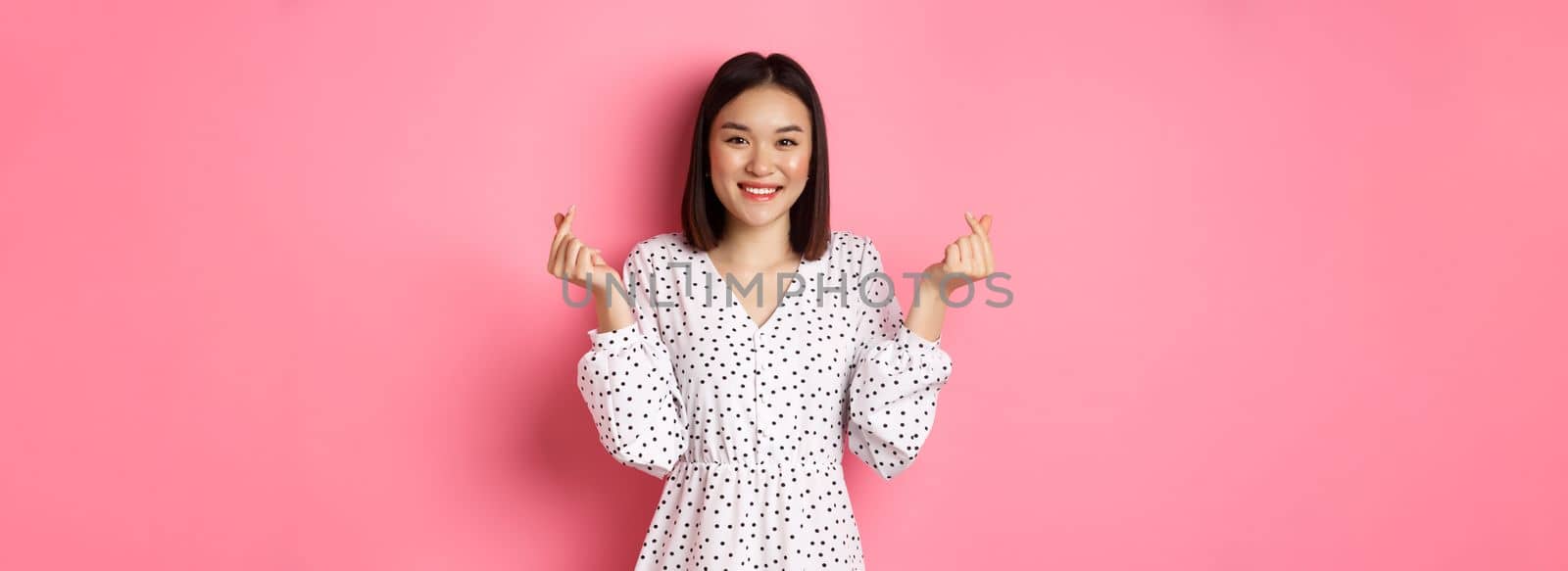 Lovely asian woman in dress showing korean heart signs and smiling, standing on romantic pink background by Benzoix