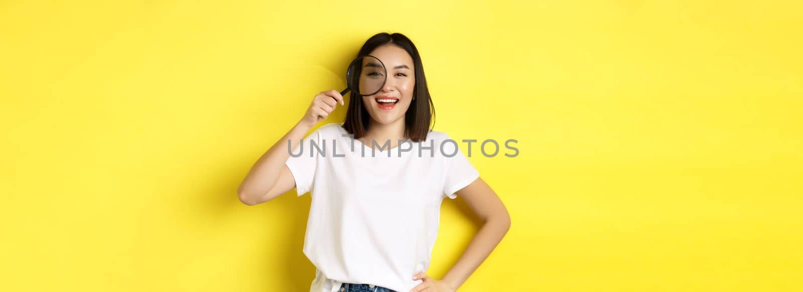 Cheerful asian girl searching for you, looking through magnifying glass and smiling, found something interesting, standing over yellow background.