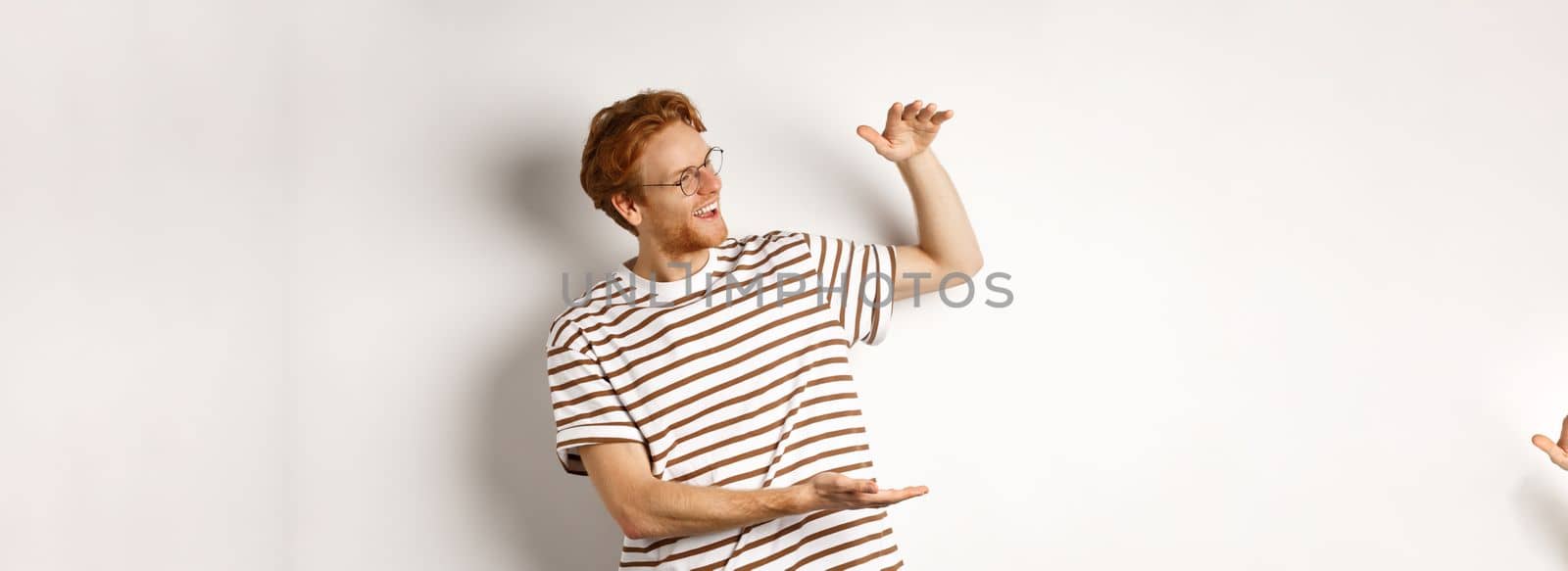 Happy and satisfied caucasian redhead man introduce something big, showing large product and smiling pleased, white background.