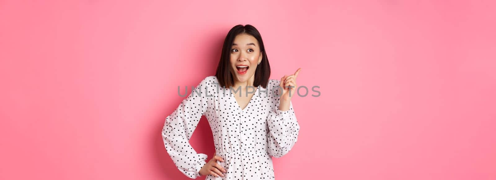 Excited asian woman in stylish dress, gasping amused, pointing finger upper left corner, showing interesting promo offer, standing over pink background.