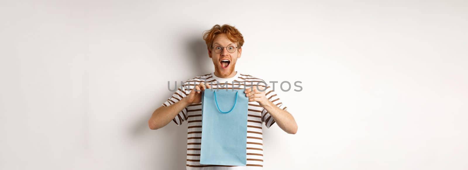 Holidays and celebration concept. Surprised redhead man receiving present inside shopping bag, looking amazed and thankful at camera, white background by Benzoix