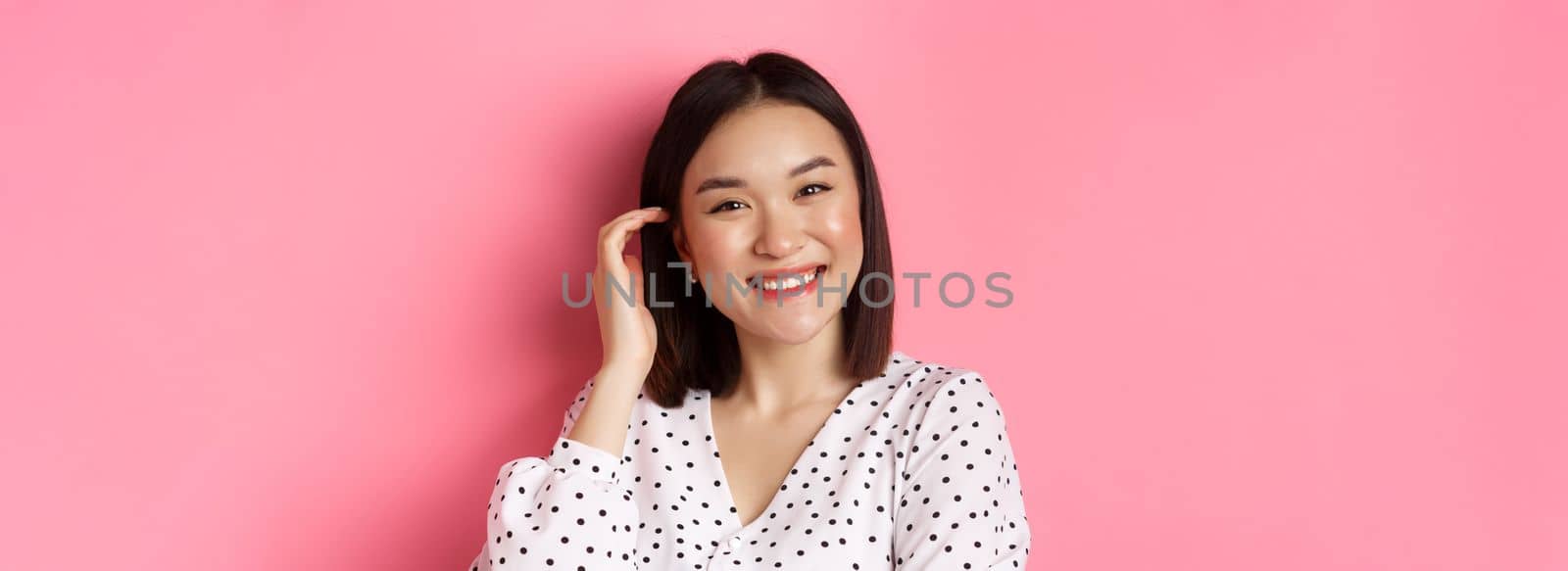 Close-up of beautiful asian woman smiling happy, touching new haircut, standing over pink background.