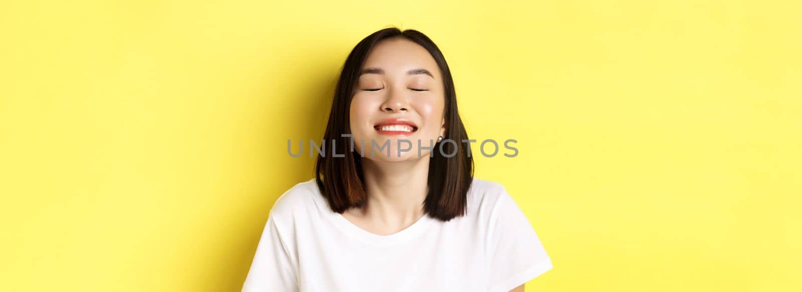 Close up of happy and relaxed asian woman enjoying sun, smiling with eyes closed and looking joyful, standing over yellow background.