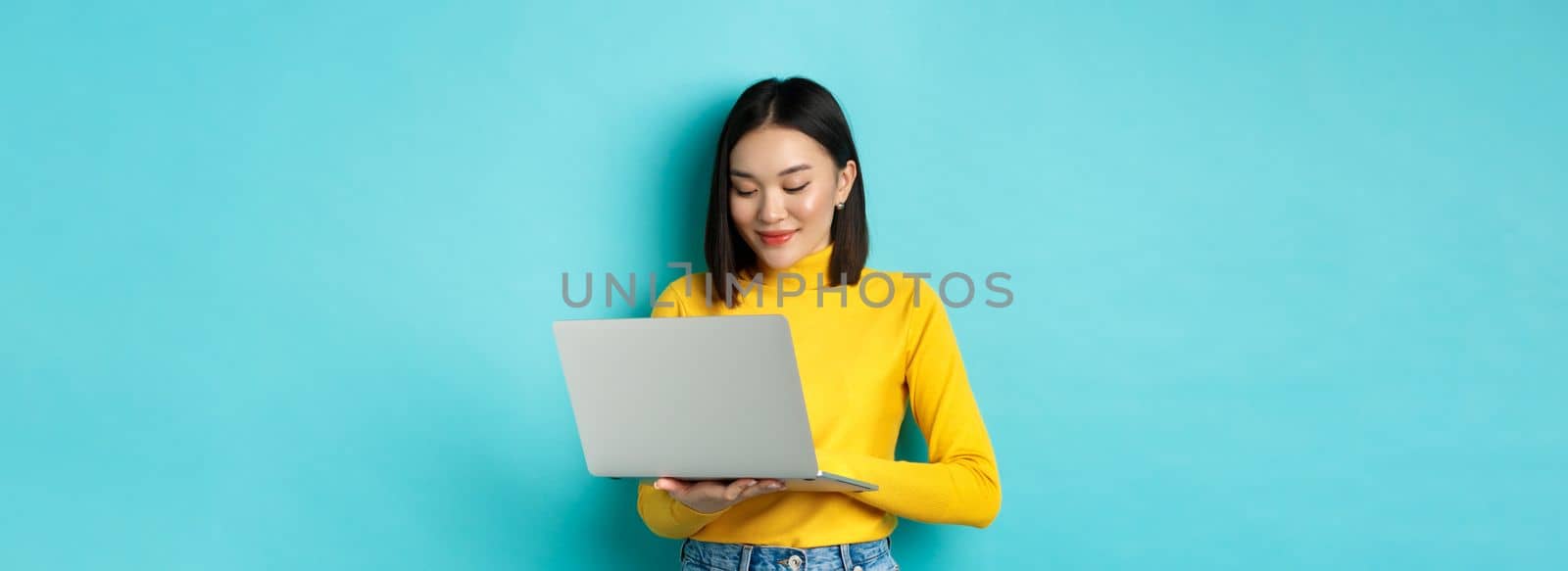 Beautiful asian female student working on laptop, typing on keyboard and looking at screen with pleased smile, standing over blue background.