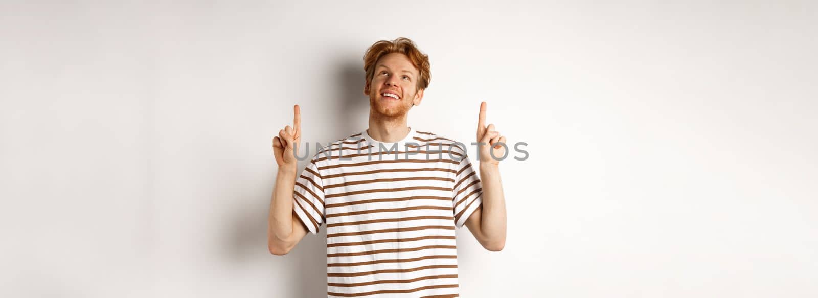 Happy young man with red hair and beard pointing, looking up with dreamy smile, checking out special promo offer, white background.