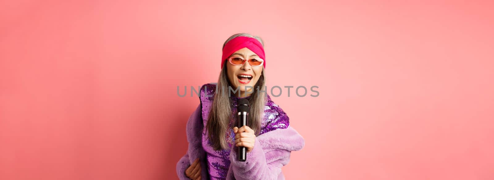 Fashion and lifestyle concept. Beautiful middle-aged woman in sunglasses, party dress and faux fur coat, singing in microphone and having fun at karaoke bar, pink background.