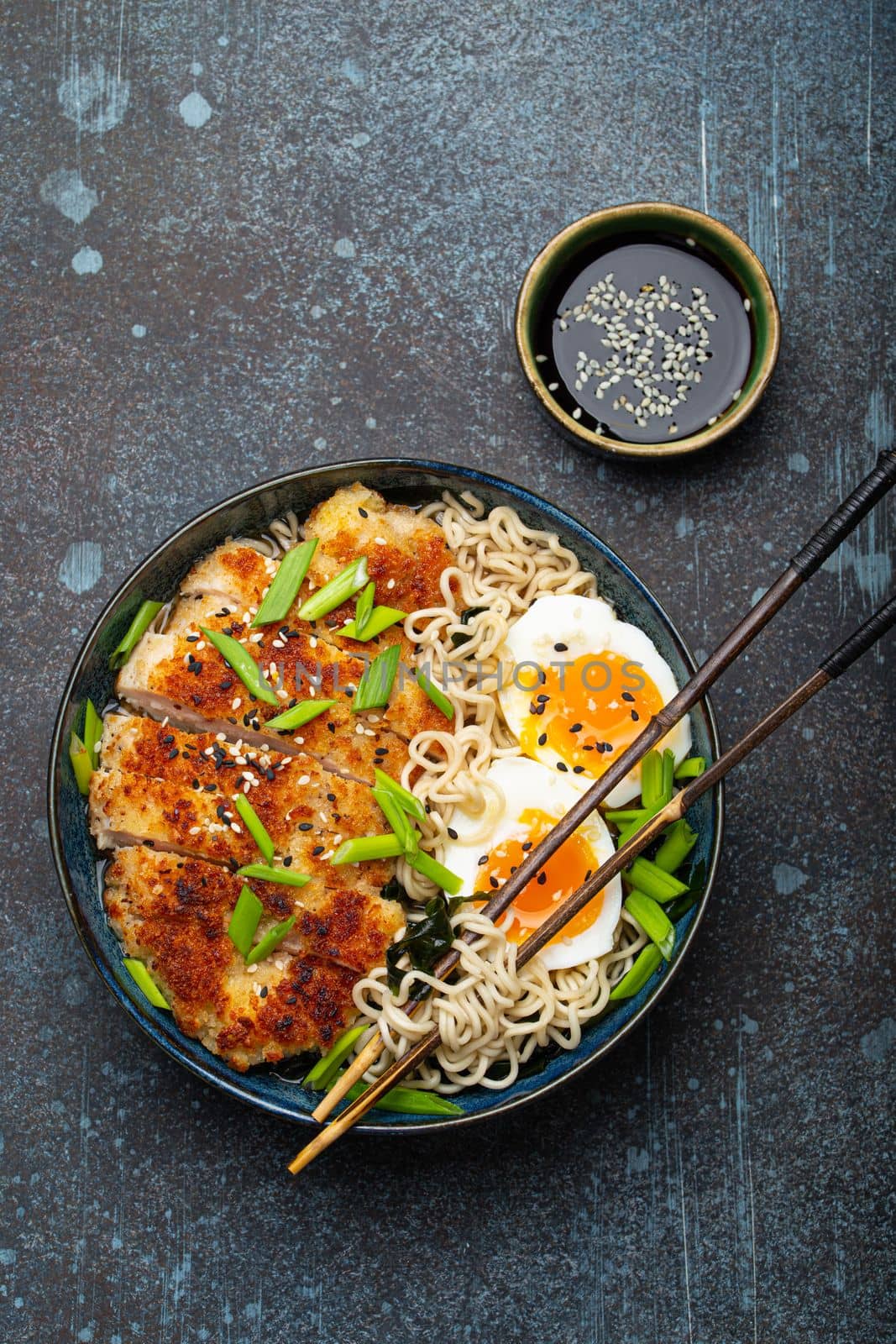 Asian noodles ramen soup with deep fried panko chicken fillet and boiled eggs in ceramic bowl with chop sticks, soy sauce on stone rustic background top view by its_al_dente