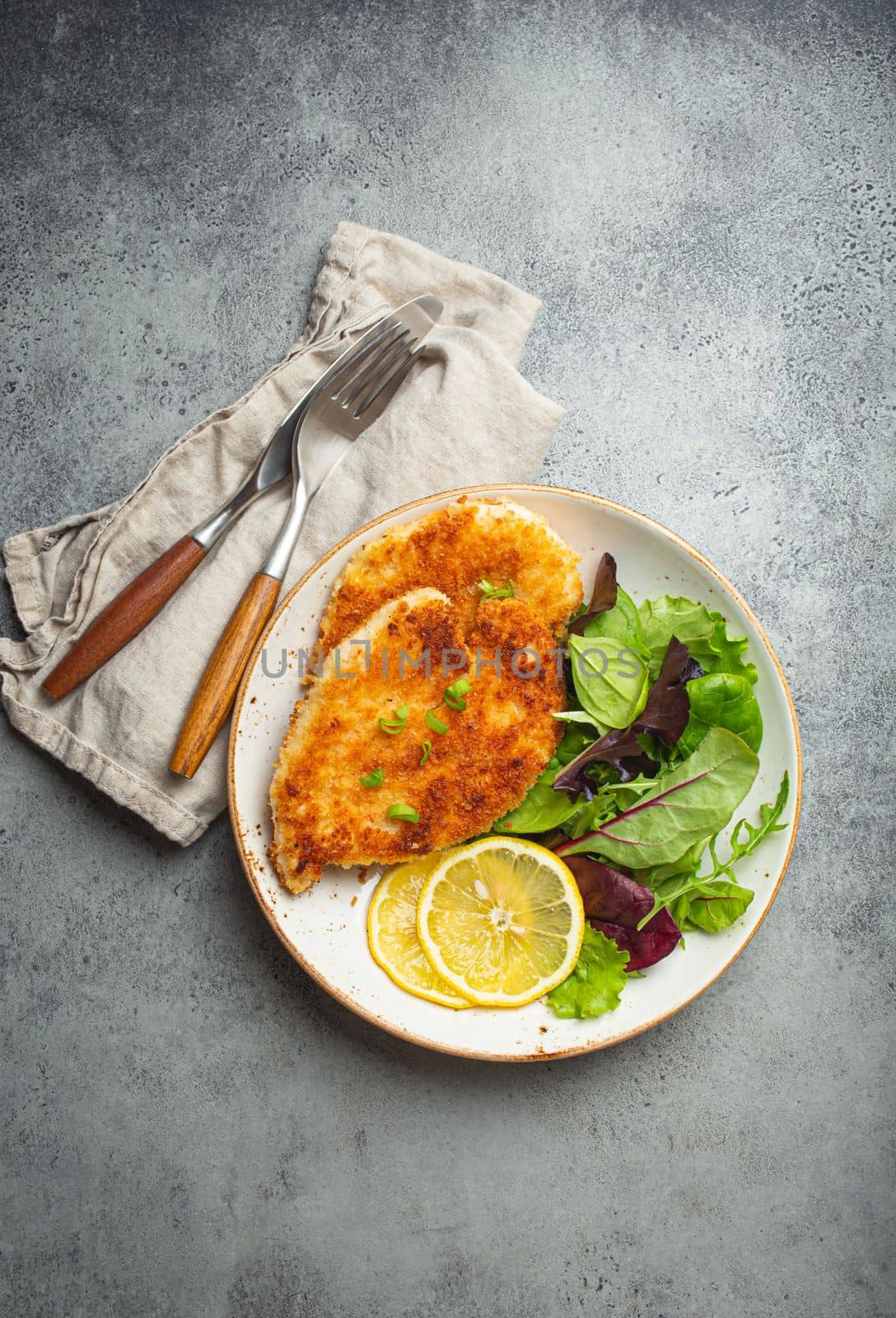Crispy panko breaded fried chicken fillet with green salad and lemon on plate with cutlery on gray rustic concrete background table from above. Japanese style deep fried coated chicken breasts .