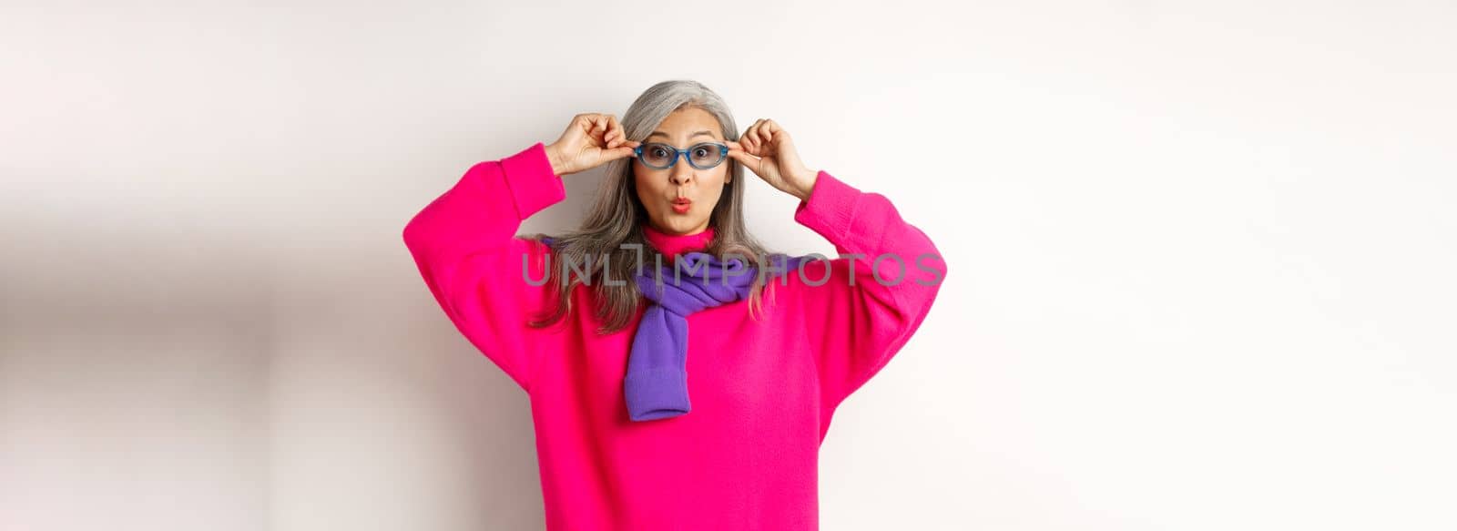 Trendy asian grandmother in sunglasses, pucker lips and looking surprised at camera, staring amazed at special offer, standing over white background.