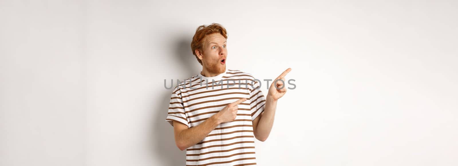 Check it out. Amazed redhead guy pointing and looking left at promotion banner, saying wow, standing over white background.