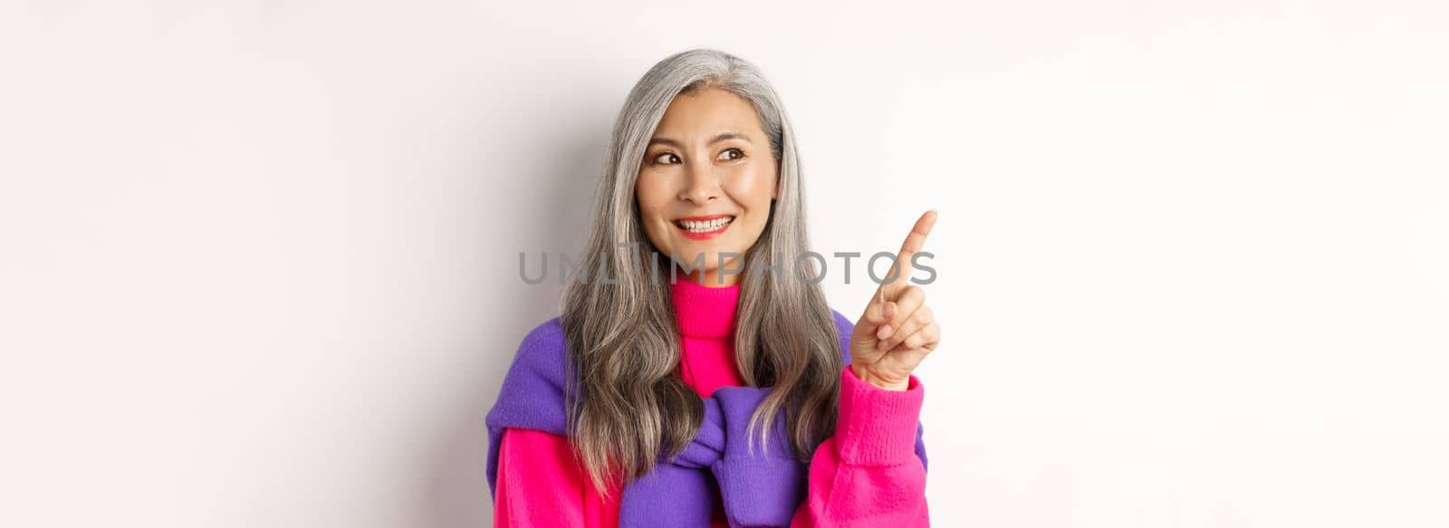 Close-up of beautiful asian senior woman smiling, looking and pointing upper left corner advertisement, checking out promo offer, standing over white background by Benzoix