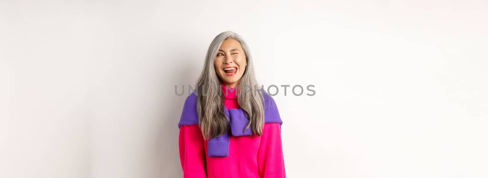 Funny and happy asian senior woman having fun, showing tongue and winking cheeky, looking left at logo, standing over white background.