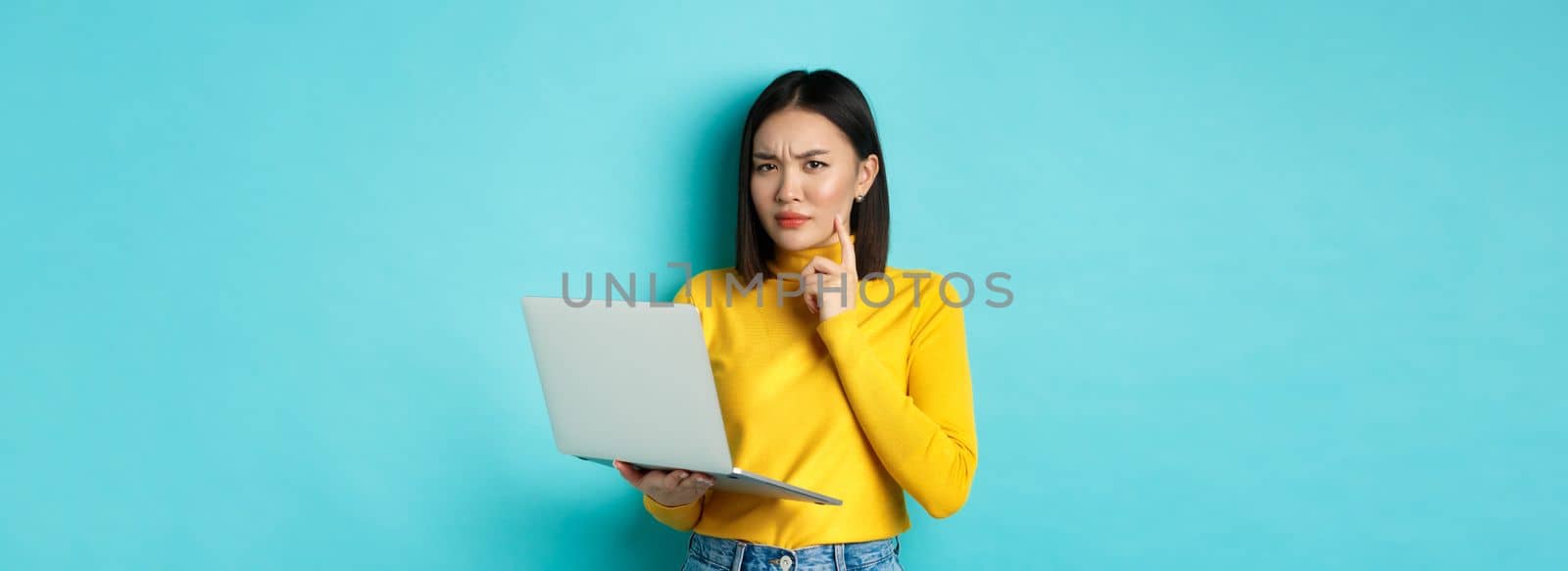 Serious looking asian woman working on laptop and thinking, frowning at camera, solving problem at work, standing over blue background.