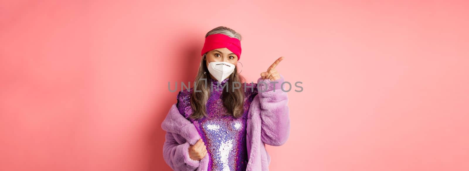 Covid and fashion concept. Fashionable asian mature woman in face mask and disco dress pointing finger upper left corner. Old lady in glittering dress showing advertisement.