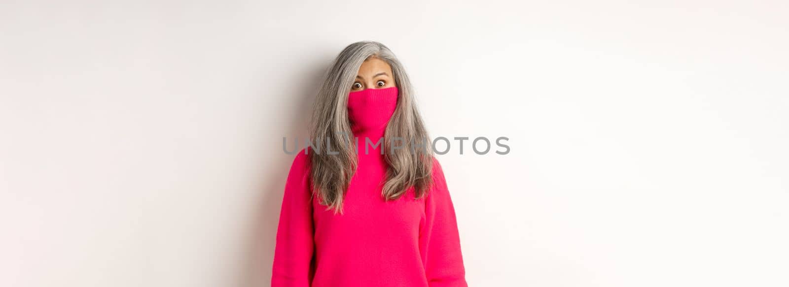 Funny and cute asian senior woman pull sweater collar on face and looking at camera, standing over white background by Benzoix