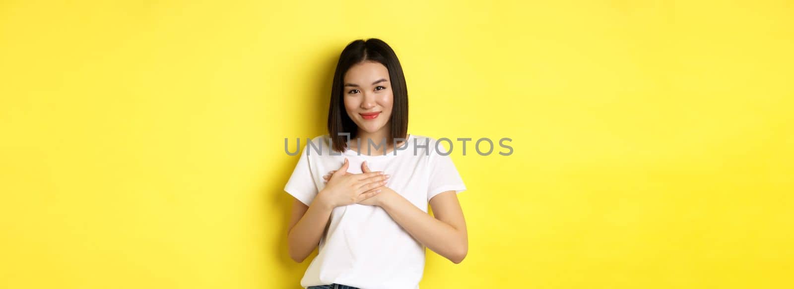 Beauty and fashion concept. Heartfelt asian girl holding hands on heart and smiling touched, thanking you, standing over yellow background.