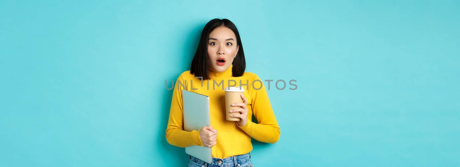 Shocked asian woman employee, drinking coffee from cafe takeaway, holding laptop, gasping and staring amazed at camera, standing over blue background.