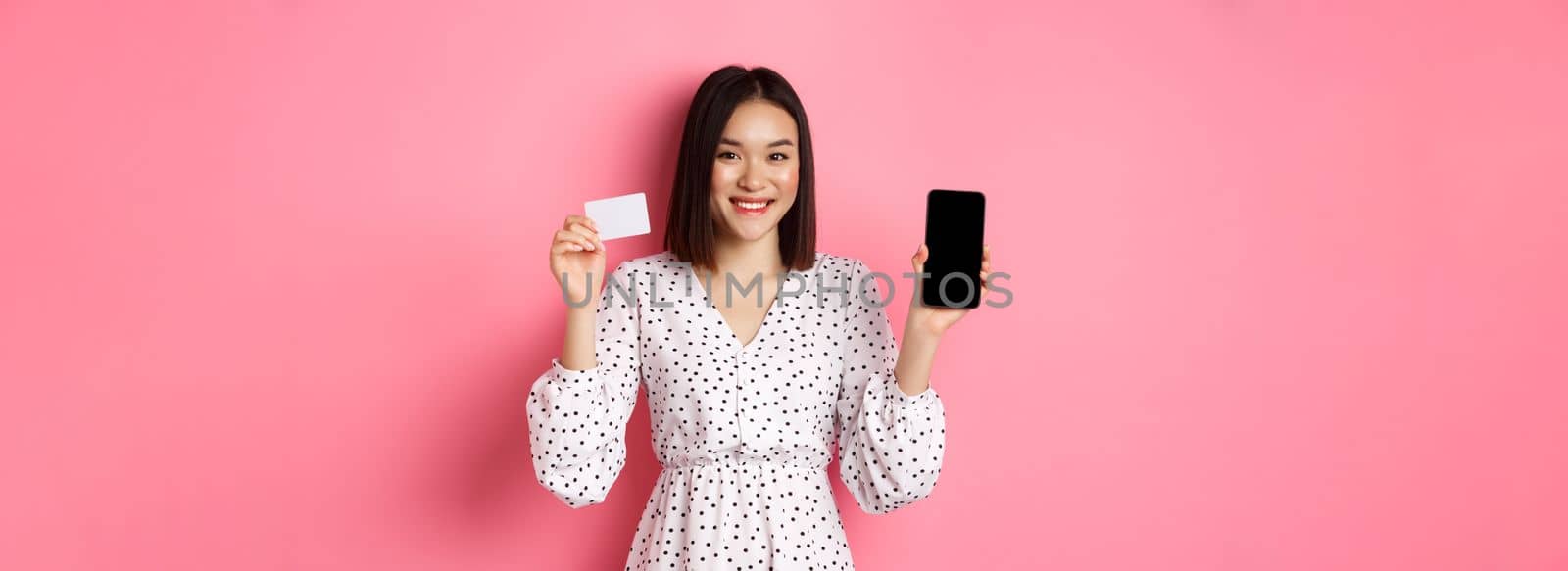 Cute asian woman shopping online, showing bank credit card and mobile screen, smiling and looking at camera, standing over pink background by Benzoix