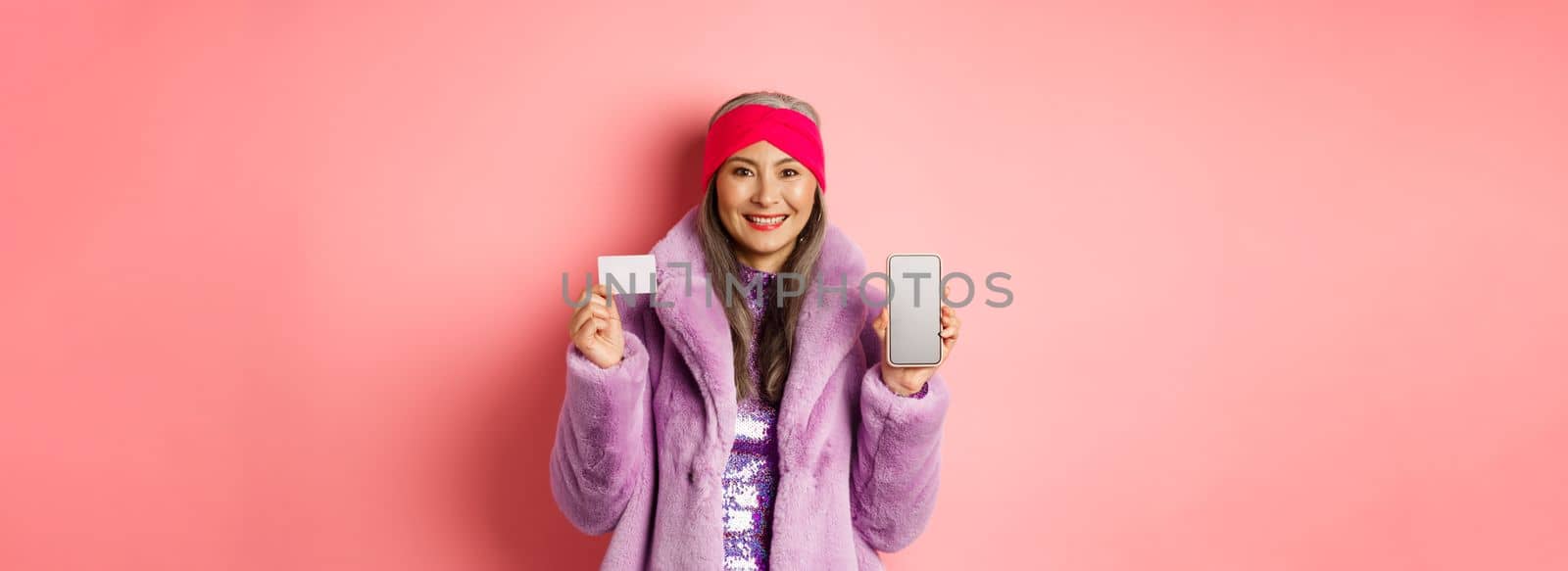 Online shopping and fashion concept. Fashionable asian senior woman showing black screen of smartphone and plastic credit card, smiling happy, pink background by Benzoix