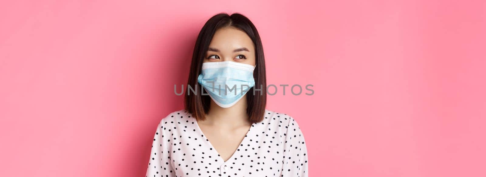 Covid-19, pandemic and lifestyle concept. Beautiful asian female model in medical mask laughing, smiling and looking left at copy space, standing over pink background by Benzoix